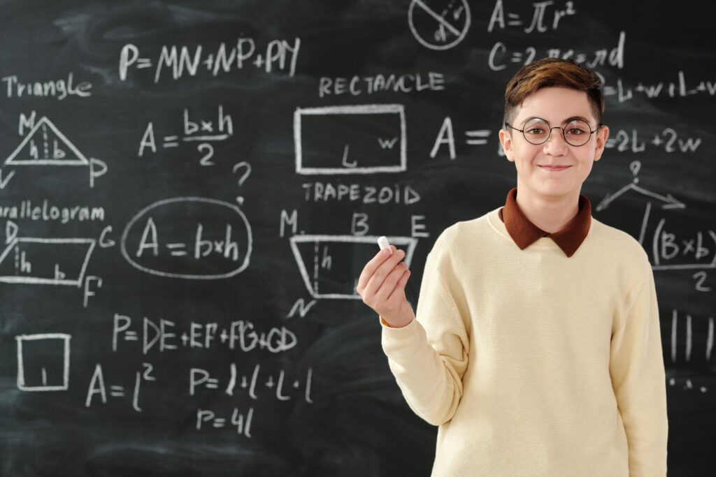 Young boy holding chalk in front of blackboard with math formulas, wearing glasses and smiling.